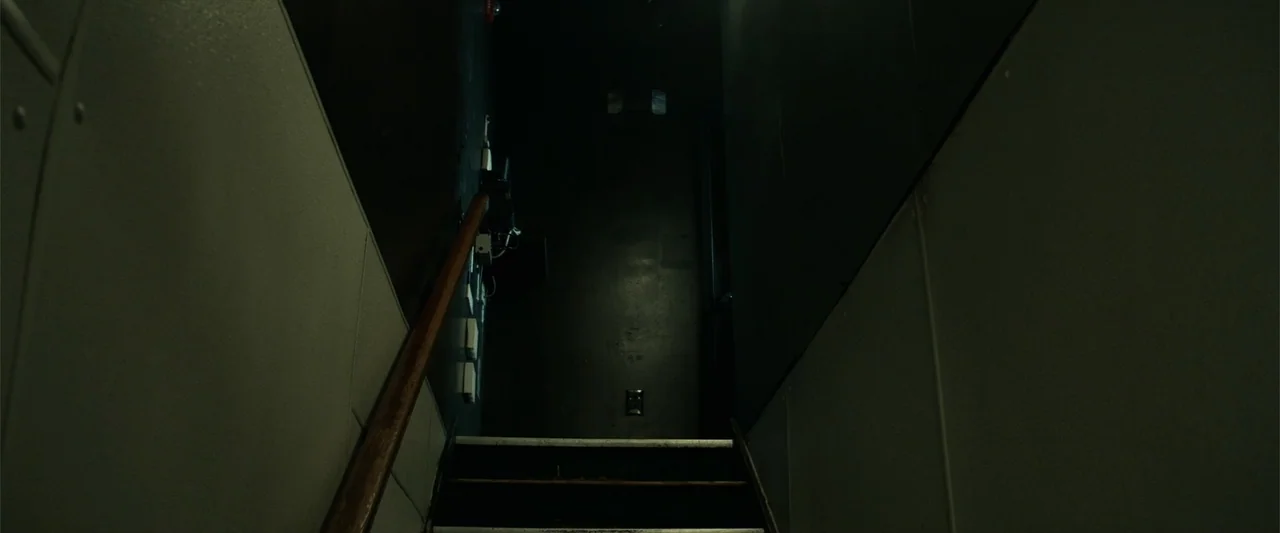 21 Bridges (2019) movie still: establishing — A dimly lit industrial stairwell features gray tiled walls and a dark wooden handrail. The perspective…