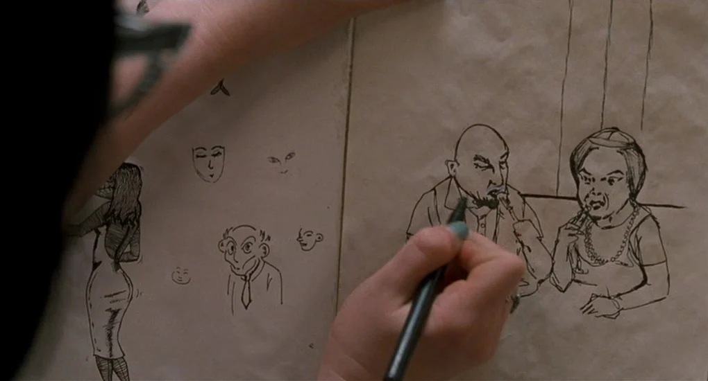 Ghost World (2001) movie still: closeup — A sketchpad rests on a surface, covered with hand-drawn ink caricatures of people. A person's hands…
