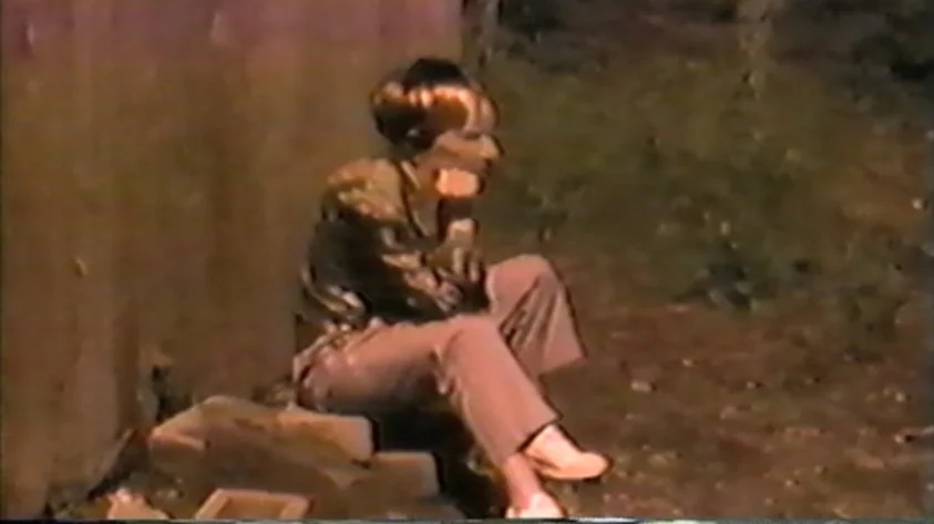 Trash Humpers (2009) movie still: fullbody — A person sits on a makeshift stack of wooden blocks against a weathered wall. The area is dark and…