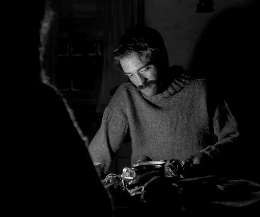 The Lighthouse (2019) movie still: medium — A dark dining interior with a man sitting at a wooden table. The man faces across the table toward an…