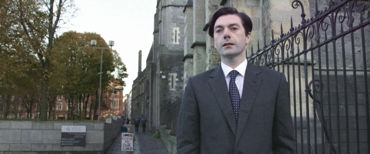 The Cured (2017) movie still: medium — An outdoor area beside a large stone building with an ornamental iron fence. A man in a dark suit stands…