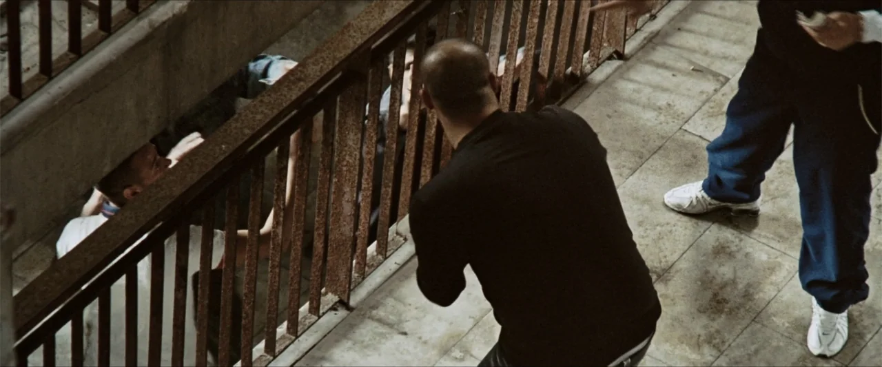 GOMORRAH (2023) movie still: medium — A concrete balcony overlooks a lower level with a rusted iron railing separator. A man in a black shirt stands…