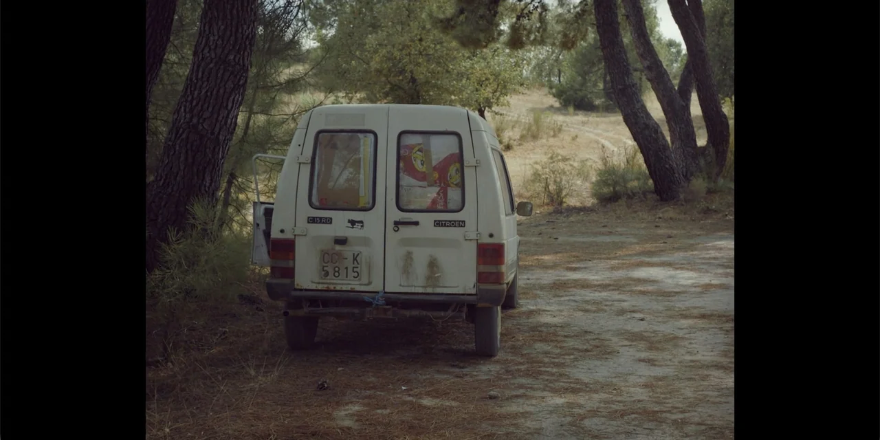 La coda del diavolo (2024) movie still: medium — A white Citroen C15 van is parked on a dirt path between pine trees. The rear doors of the vehicle…