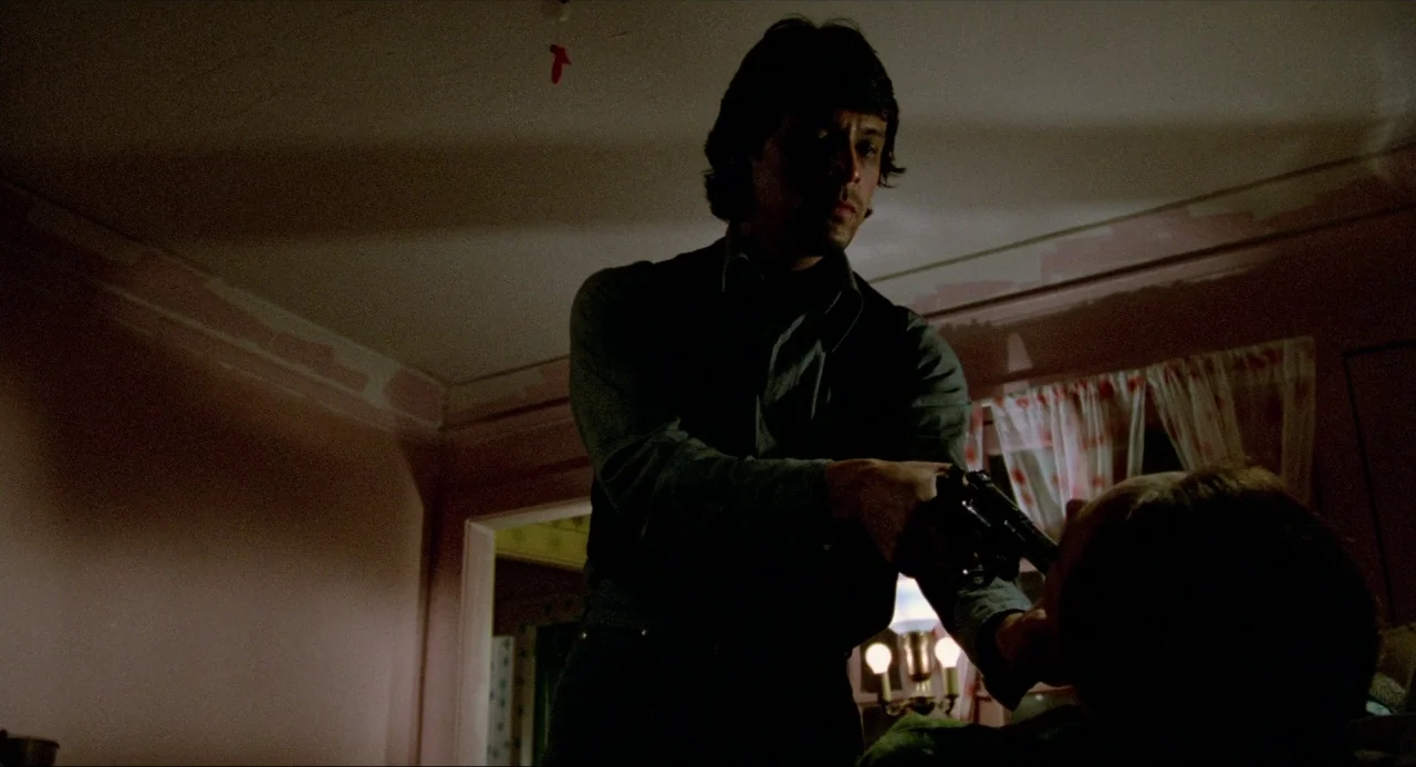 The Driver (1978) movie still: medium — A dimly lit room with muted pink walls and a window covered by curtains. A man points a handgun at a person…