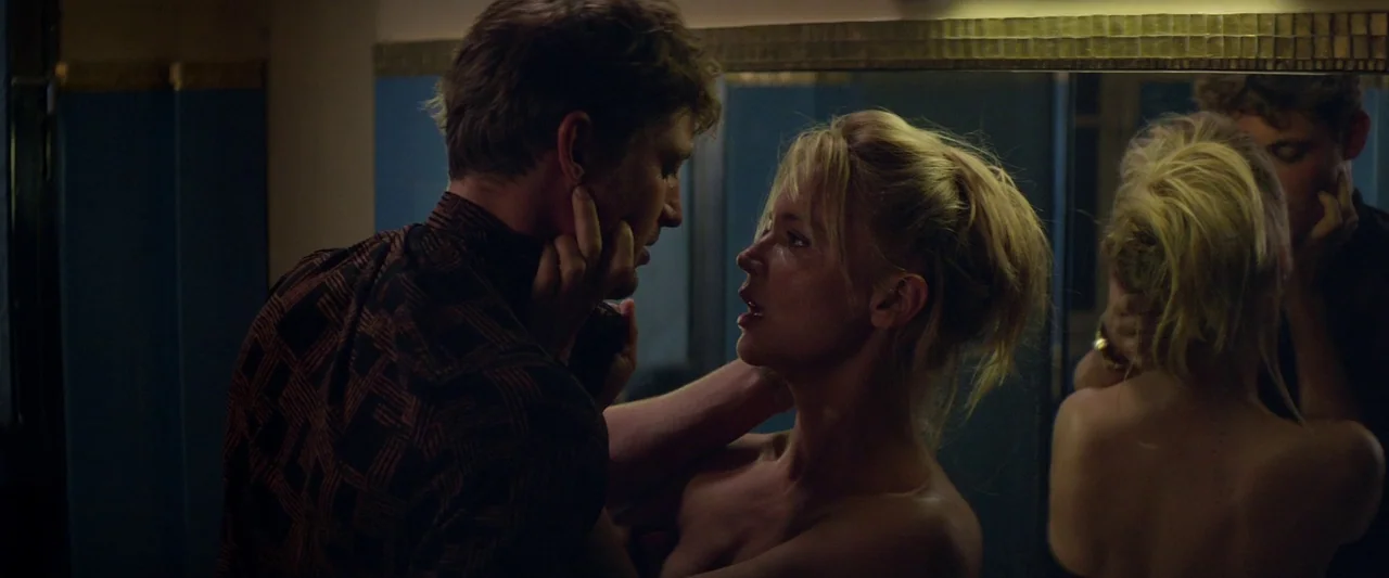 Sibyl (2019) movie still: medium — A dimly lit room with dark blue wall tiling and a reflective mirror. A man and a woman stand close together while…
