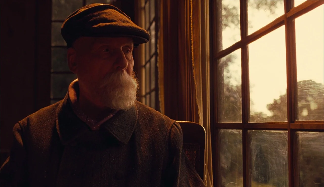 Renoir (2012) movie still: medium — A dimly lit room with wood-paneled walls features a large paned window at side. An elderly man in a tweed cap…