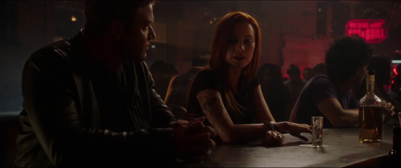 Jigsaw (2017) movie still: medium — A dimly lit bar at night with red neon signs glowing in the background. A man and a woman sit side-by-side at…
