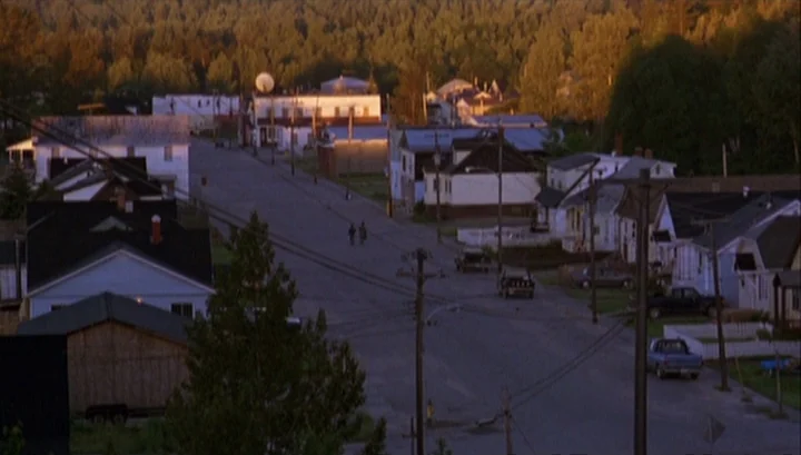 Highway 61 (1991) movie still: establishing — A quiet residential street in a small town sits nestled against a dense forest. The setting sun casts…