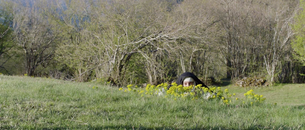 The Little Hours (2017) movie still: wide — A green grassy field with a line of bare trees in the background. A nun in a habit crouches behind a…