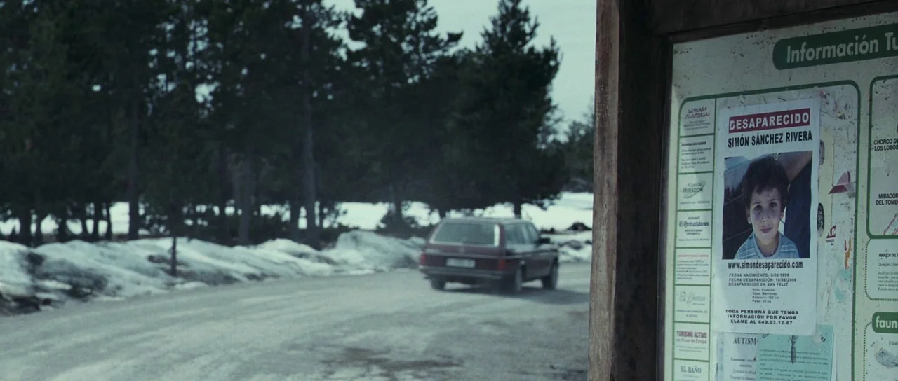 The Orphanage (2007) movie still: wide — A wooden public information board stands in the foreground, displaying a missing child poster. A maroon…