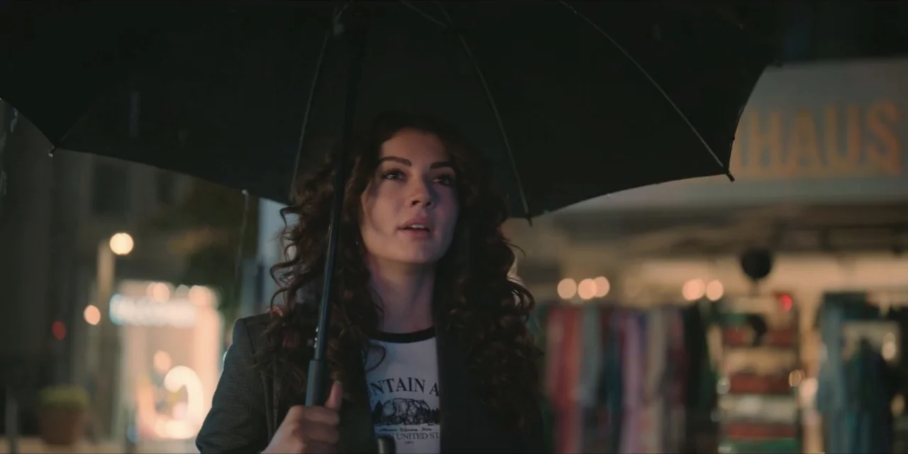Kill Me If You Dare (2019) movie still: medium — A woman stands outside on a city sidewalk beneath a large black umbrella. Soft urban lights are…