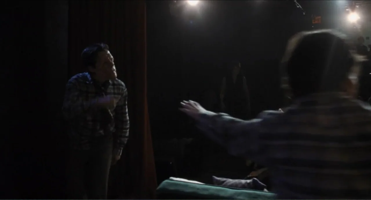 A Different Man (2024) movie still: medium — A dark indoor theater stage with a heavy red curtain hanging on the side. A man stands in the light and…