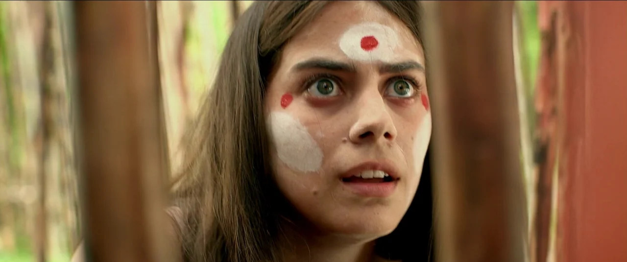 Green Inferno (2013) movie still: closeup — A young woman stands behind thin vertical bars in an outdoor setting. She peers through the obstruction…