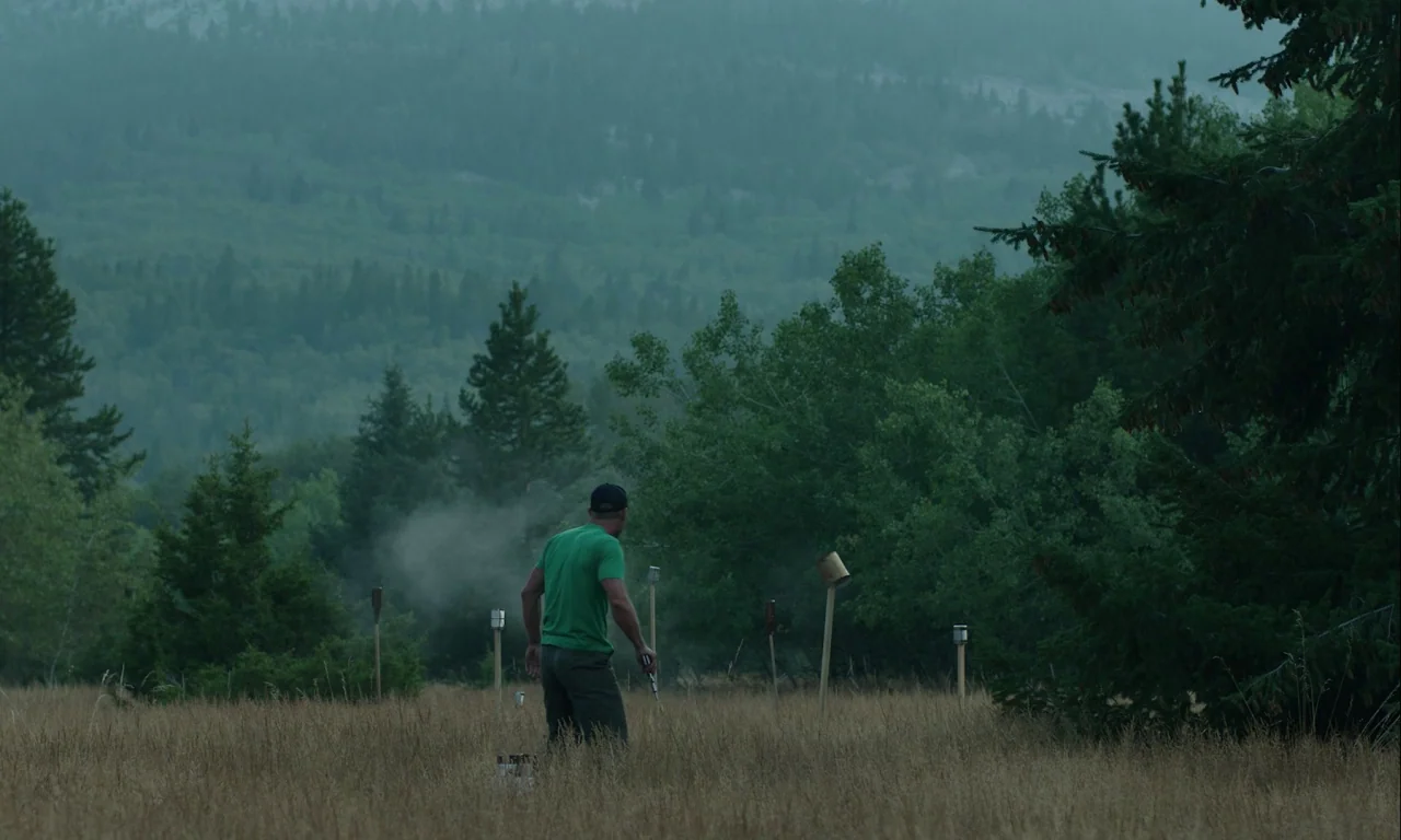 Mickey and the Bear (2019) movie still: wide — A field of dry brown grass sits before a dense treeline and mountain backdrop. A man wearing a green…