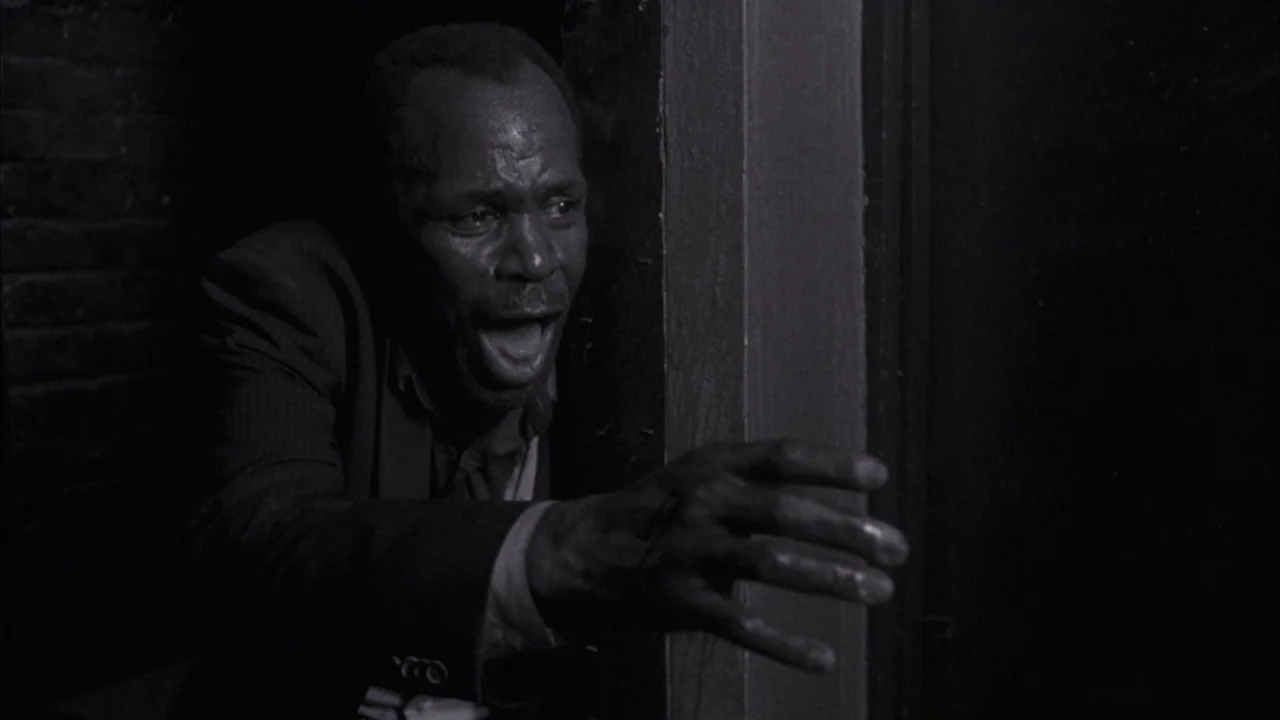 Saw movie still: medium — A dark, narrow doorway frames a man illuminated by a sharp side light. The man reaches out with an open hand and an…