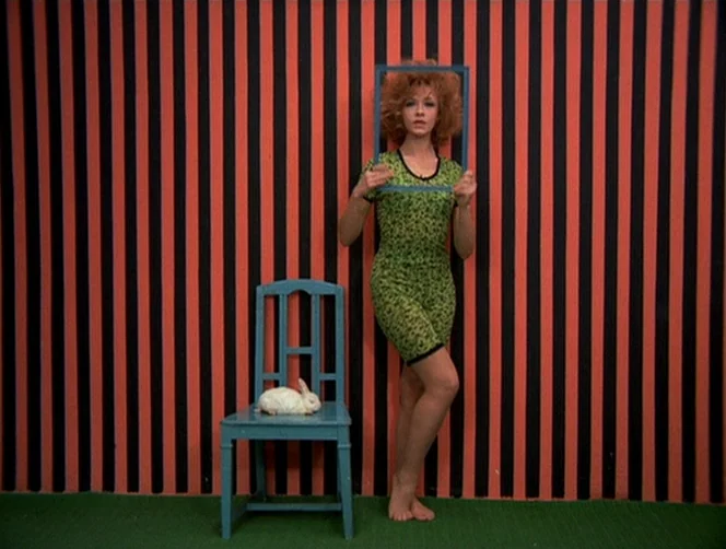 WR: Mysteries of the Organism (1971) movie still: fullbody — A woman stands barefoot in front of a wall with bold black and orange stripes. A white…