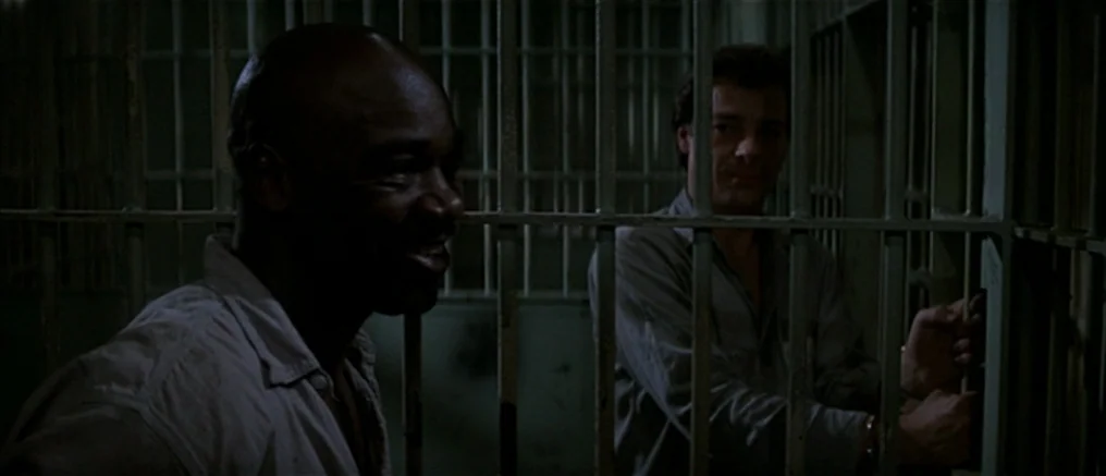 Assault on Precinct 13 (1976) movie still: medium — A dimly lit interior with heavy vertical iron bars separating the space. Two men in uniform…