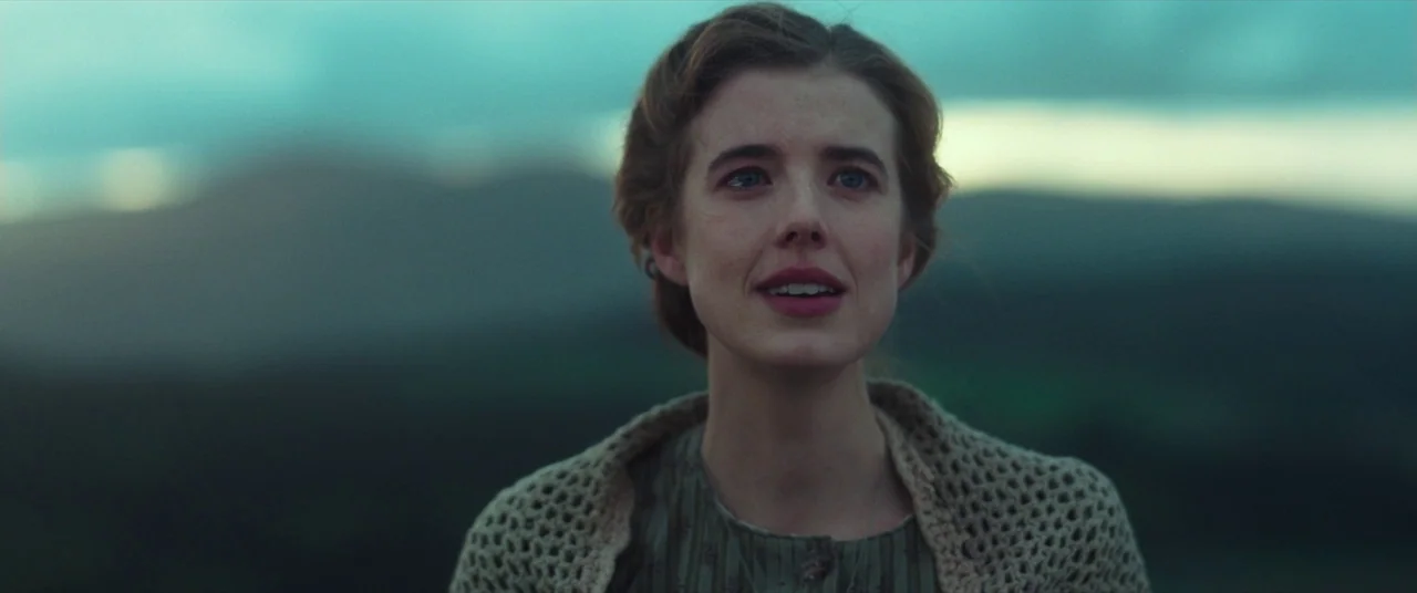 Sunset Song (2015) movie still: closeup — A woman stands in a landscape with rolling hills during a muted sunset. She looks forward with an open…