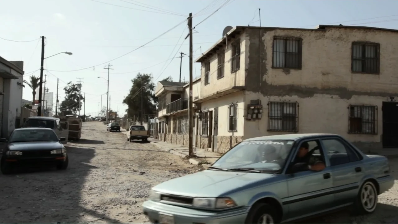 Savageland (2015) movie still: wide — A dusty residential street in a desert climate features weathered concrete houses with metal grates. A man…