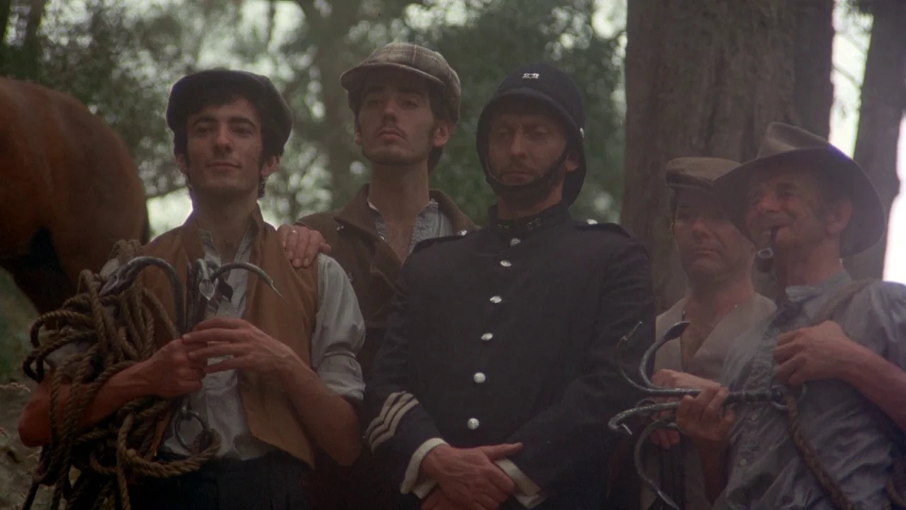 Picnic at Hanging Rock (1975) movie still: medium — A group of five men stand among trees in a dense, leafy woodland setting. The subjects hold…