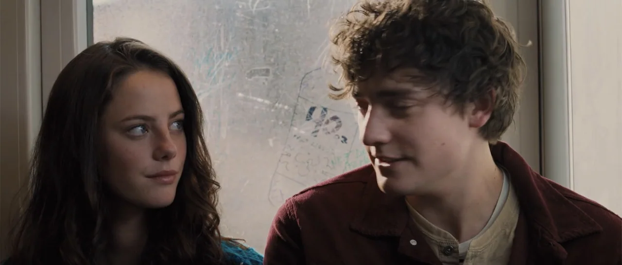 The Truth About Emanuel (2013) movie still: medium — Two young adults sit near a scratched glass window inside a public transport vehicle. They gaze…