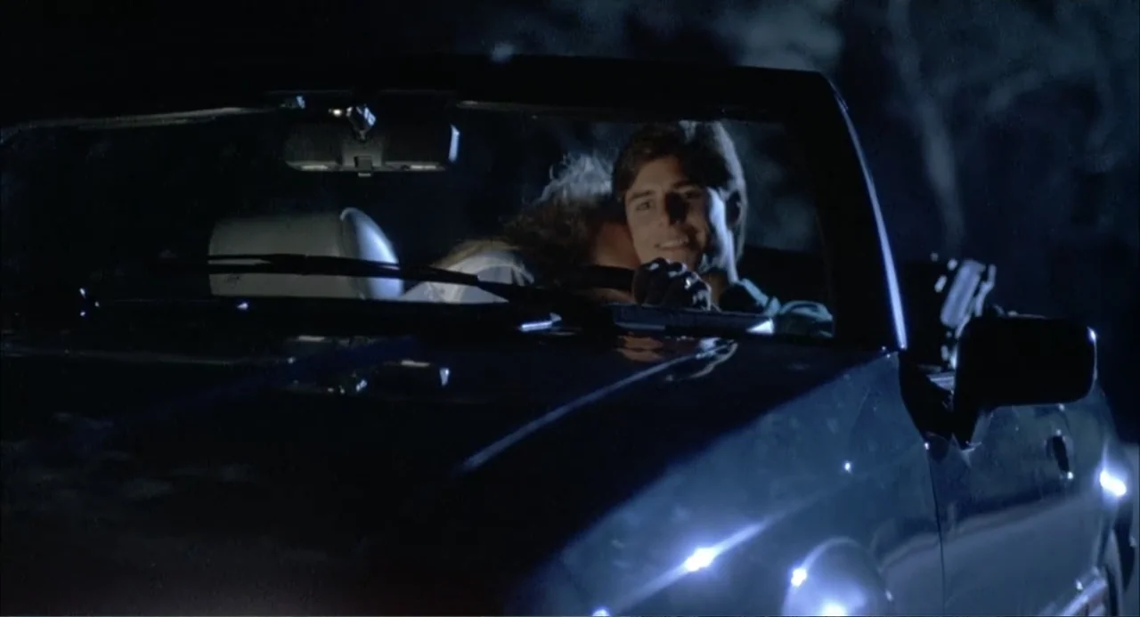 Teen Witch (1989) movie still: medium — A dark convertible drives through a night environment with the top down. A young couple sits together in the…