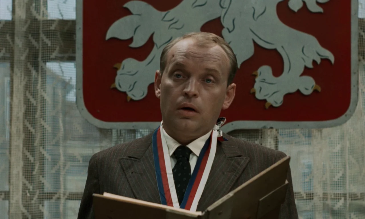 Larks On A String (1969) movie still: medium — A man in a suit stands before a wall featuring a large coat of arms. He holds a book open and speaks.