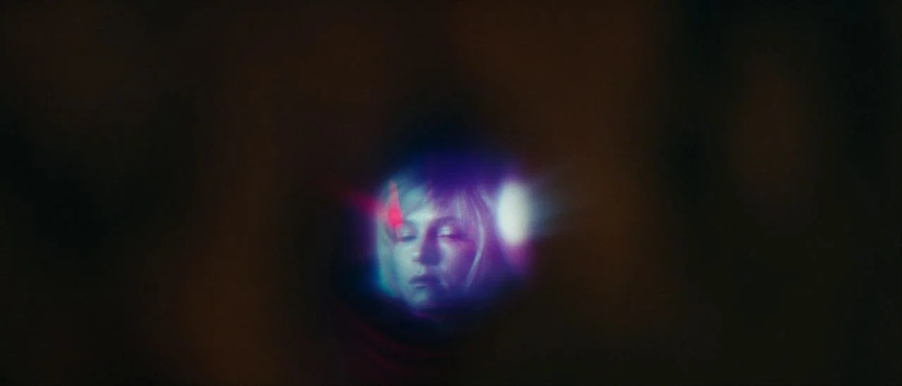 Archenemy (2020) movie still: closeup — A woman's face is viewed through a small, circular opening surrounded by darkness. Optical flares distort…