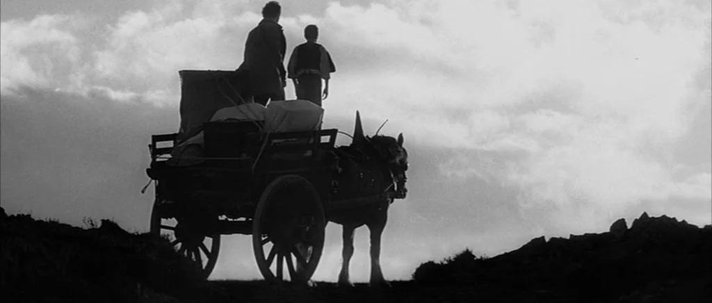 Jude (1996) movie still: wide — A rural landscape beneath a bright sky during the early 20th century. Two figures stand atop a wooden horse-drawn…