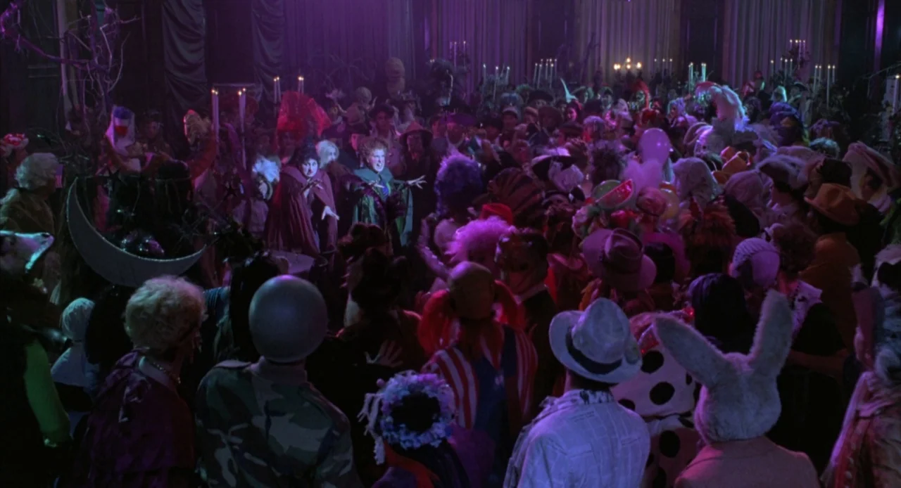 Hocus Pocus (1993) movie still: wide — A high-ceilinged room is filled with a large crowd of people wearing diverse Halloween costumes. Three women…