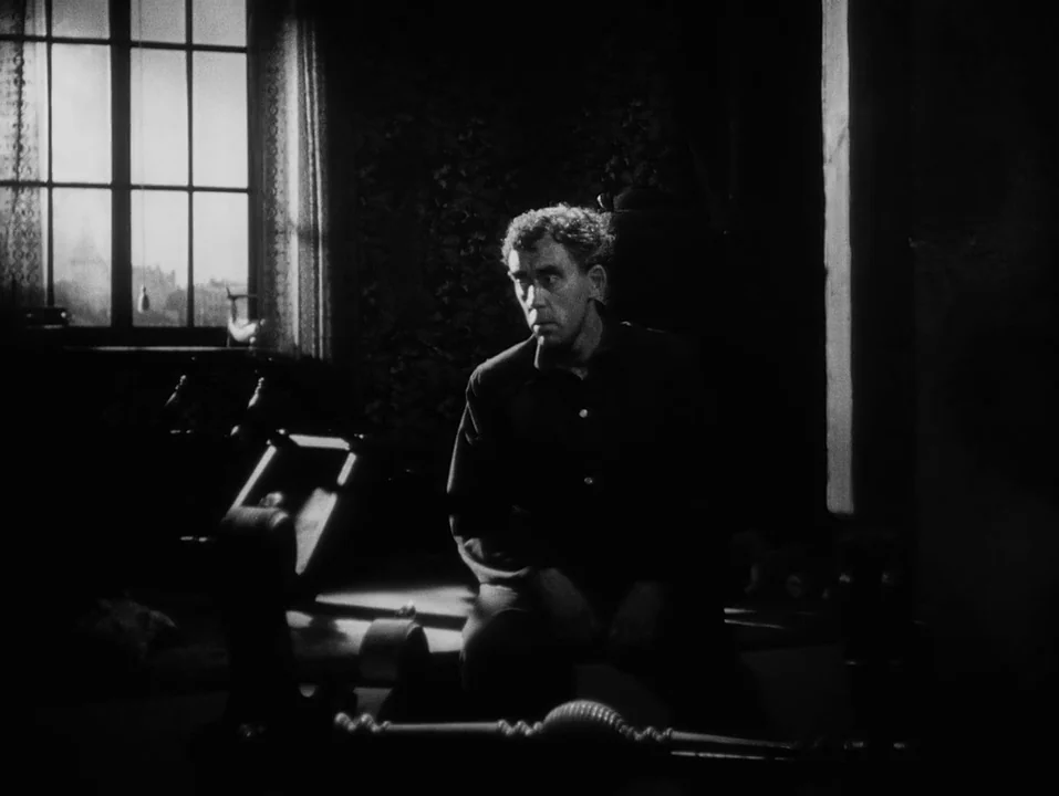 A Ship Bound for India (1947) movie still: medium — A dark, sparsely furnished room features patterned wallpaper and a large, multi-pane window. A…