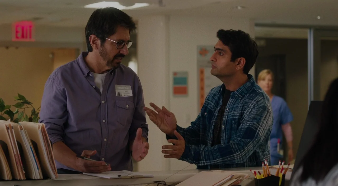 The Big Sick (2017) movie still: medium — A hospital front desk area contains a stack of files and office supplies. Two men stand together while…