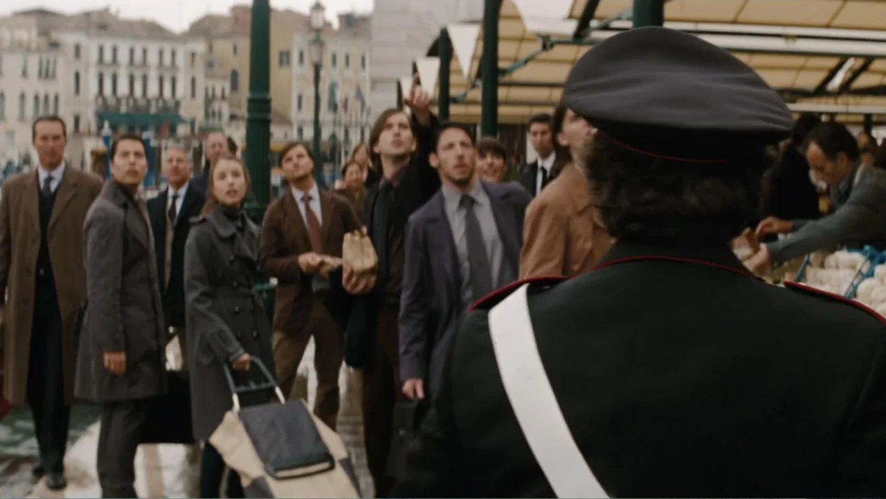 The Tourist (2010) movie still: medium — An outdoor market near a waterfront area with multiple people standing together. A police officer in…
