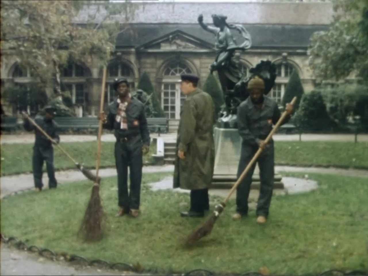 A Dessert for Constance (1981) movie still: fullbody — A public park features a central bronze statue and a stone building background. Three workers…