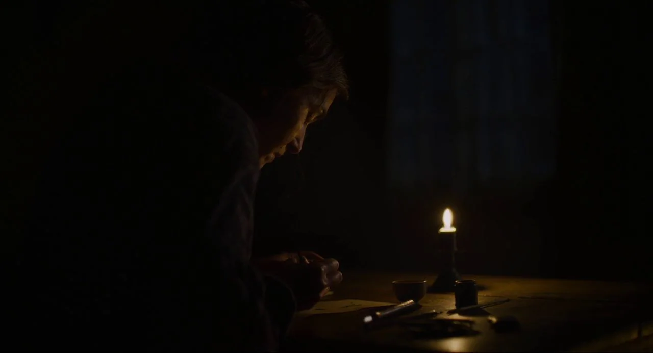 Ammonite (2020) movie still: medium — A dark, dimly lit study is illuminated by a single flickering candle on a table. A woman sits in profile…