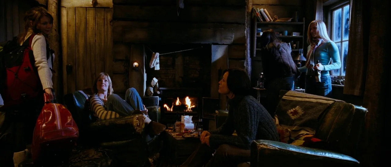 The Descent (2005) movie still: wide — A rustic wooden cabin features a large stone fireplace and leather furniture arranged centrally. Five women…