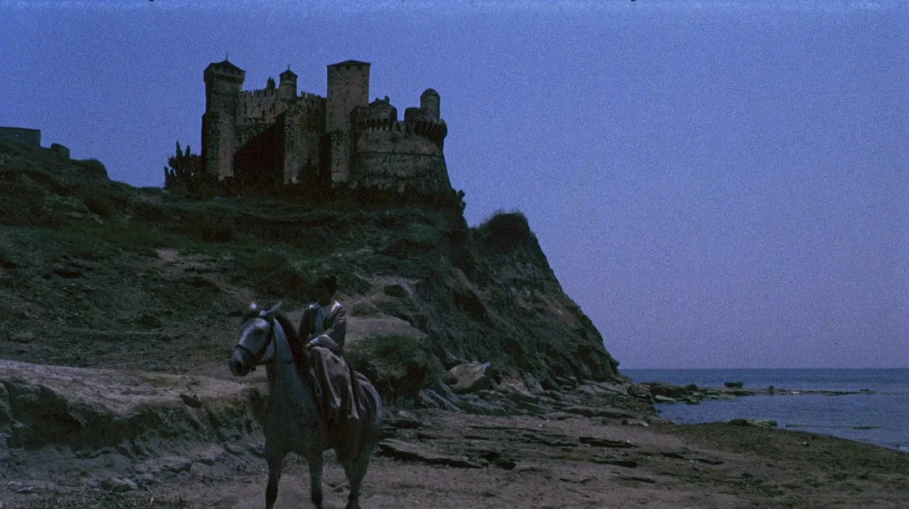 The Whip and The Body (1963) movie still: wide — A stony beach lies beneath a large stone castle perched on a rocky cliff. A woman in a historical…