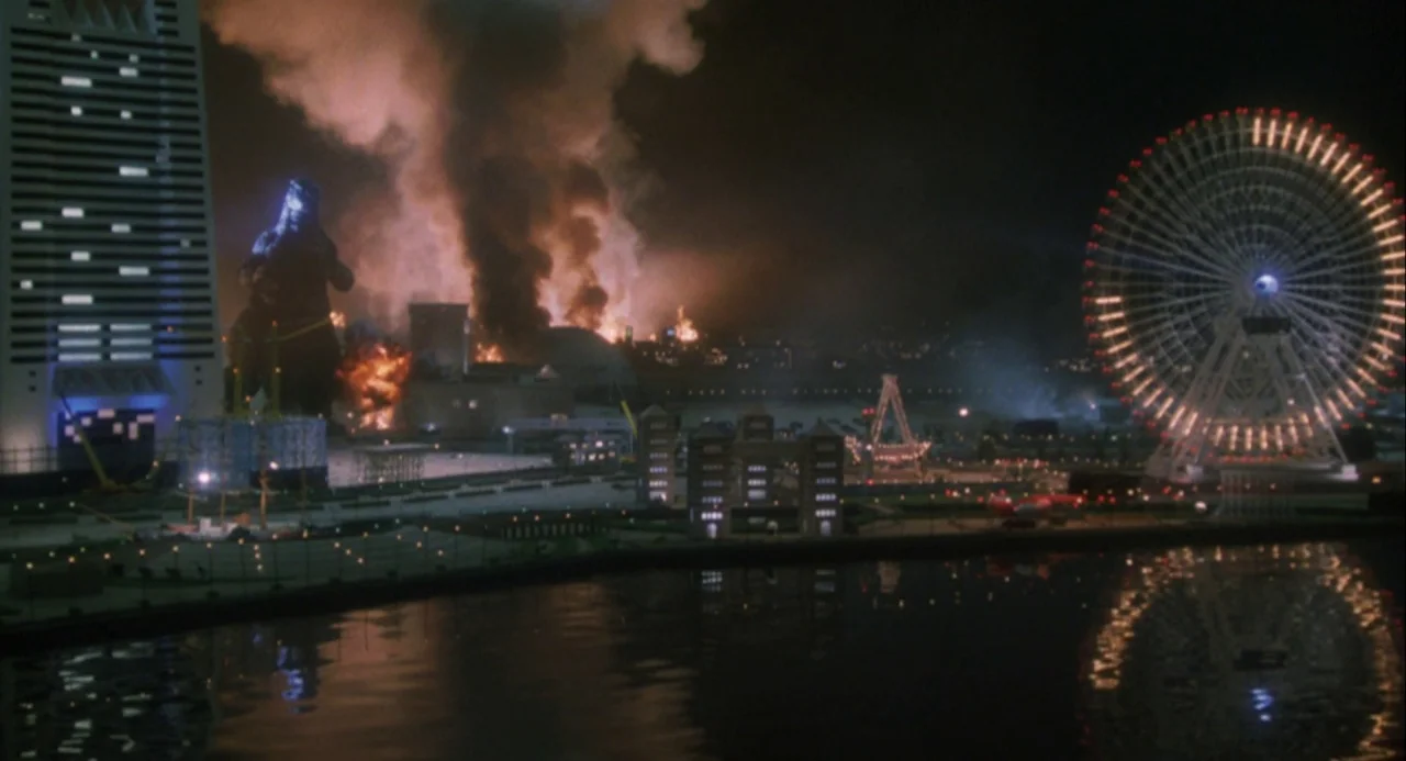 Godzilla and Mothra: The Battle for Earth (1992) movie still: wide — A nighttime city waterfront features a large burning area with black smoke…