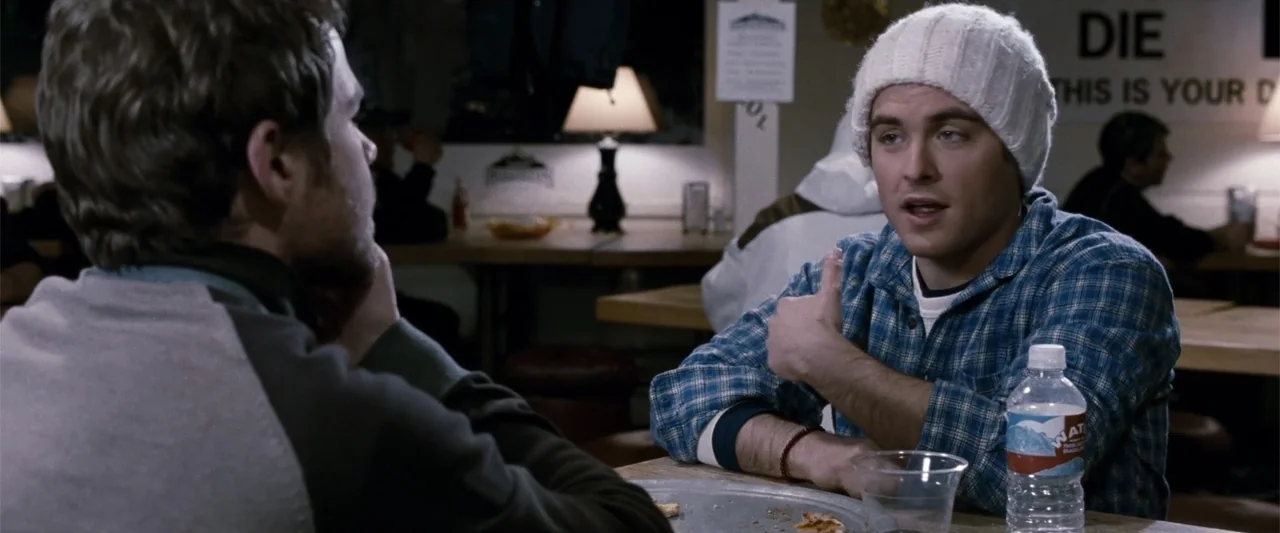 Frozen (2010) movie still: medium — A diner booth with wooden tables and a sign visible on the rear wall. A man in a blue plaid shirt talks to…