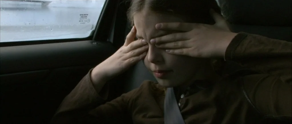 Genova (2008) movie still: closeup — The interior of a moving vehicle shows the backseat passenger window and headrest. A young child covers her…