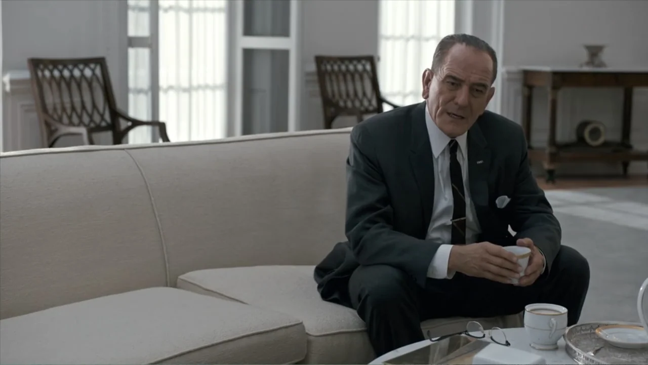 All the Way (2016) movie still: medium — A spacious and minimalist room features white walls, a curved beige sofa, and wood chairs. An older man in…