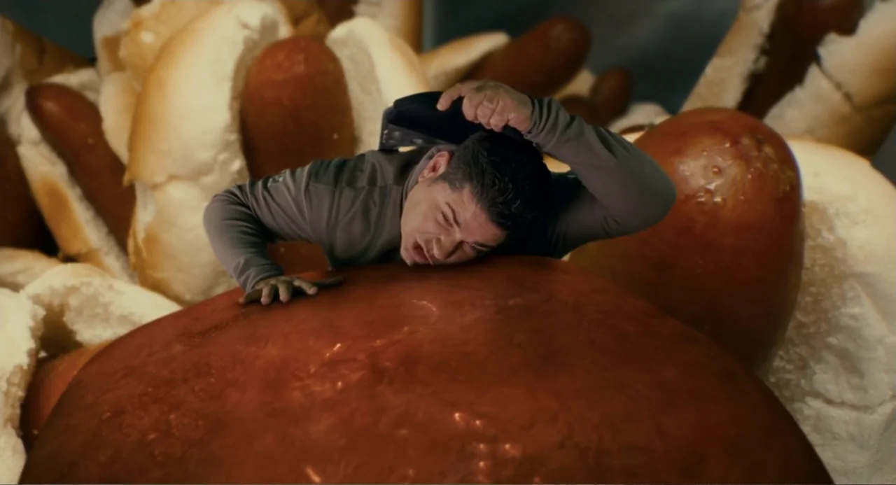 Meet Dave (2008) movie still: medium — A man is positioned in front of a giant pile of hot dogs and buns. The man presses his face against the…