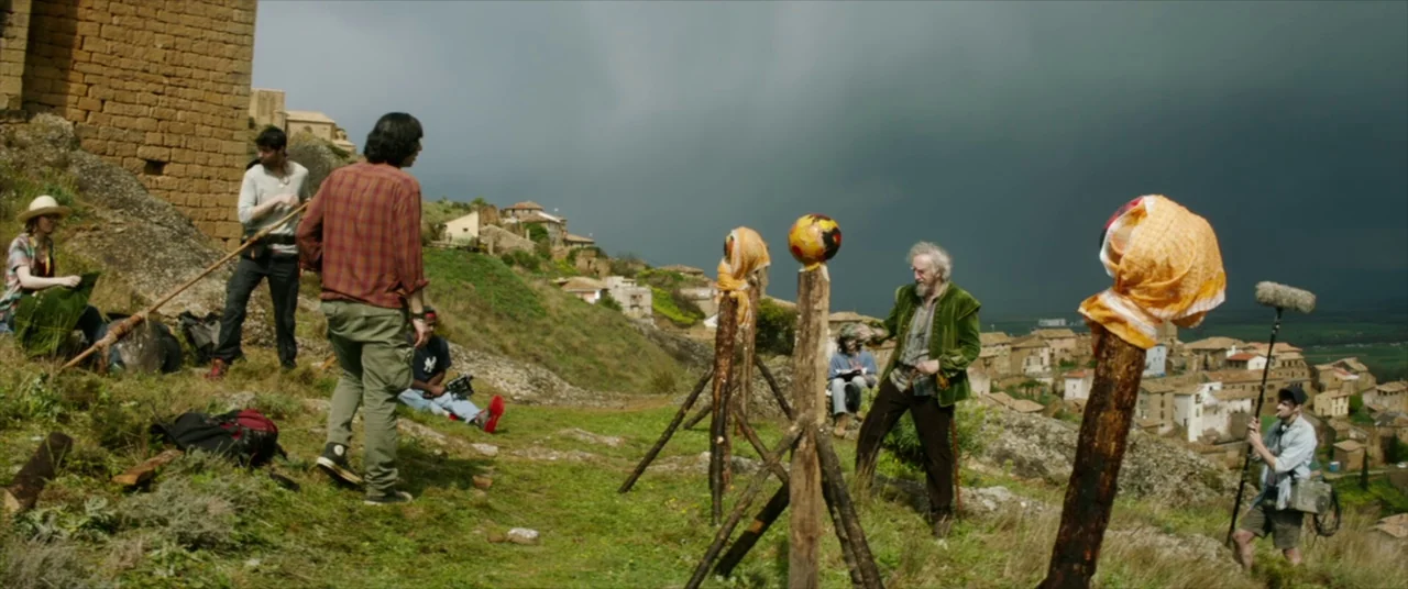 The Man Who Killed Don Quixote (2018) movie still: wide — A film crew works on a grassy hillside overlooking an old stone village. An elderly man in…