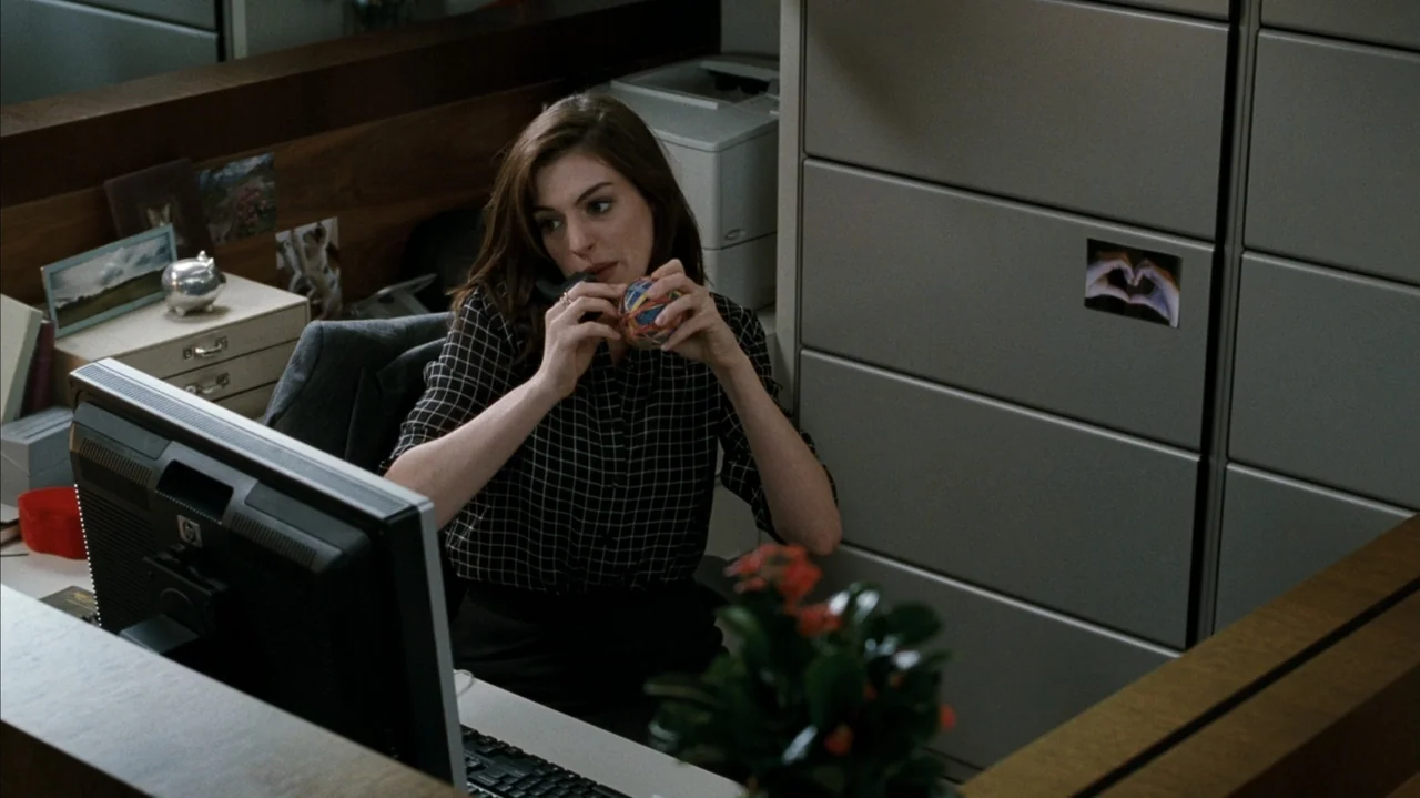 F Valentine's Day (2026) movie still: medium — A woman sits in a corporate cubicle with a desk monitor and gray partitions. She is wearing a dark…