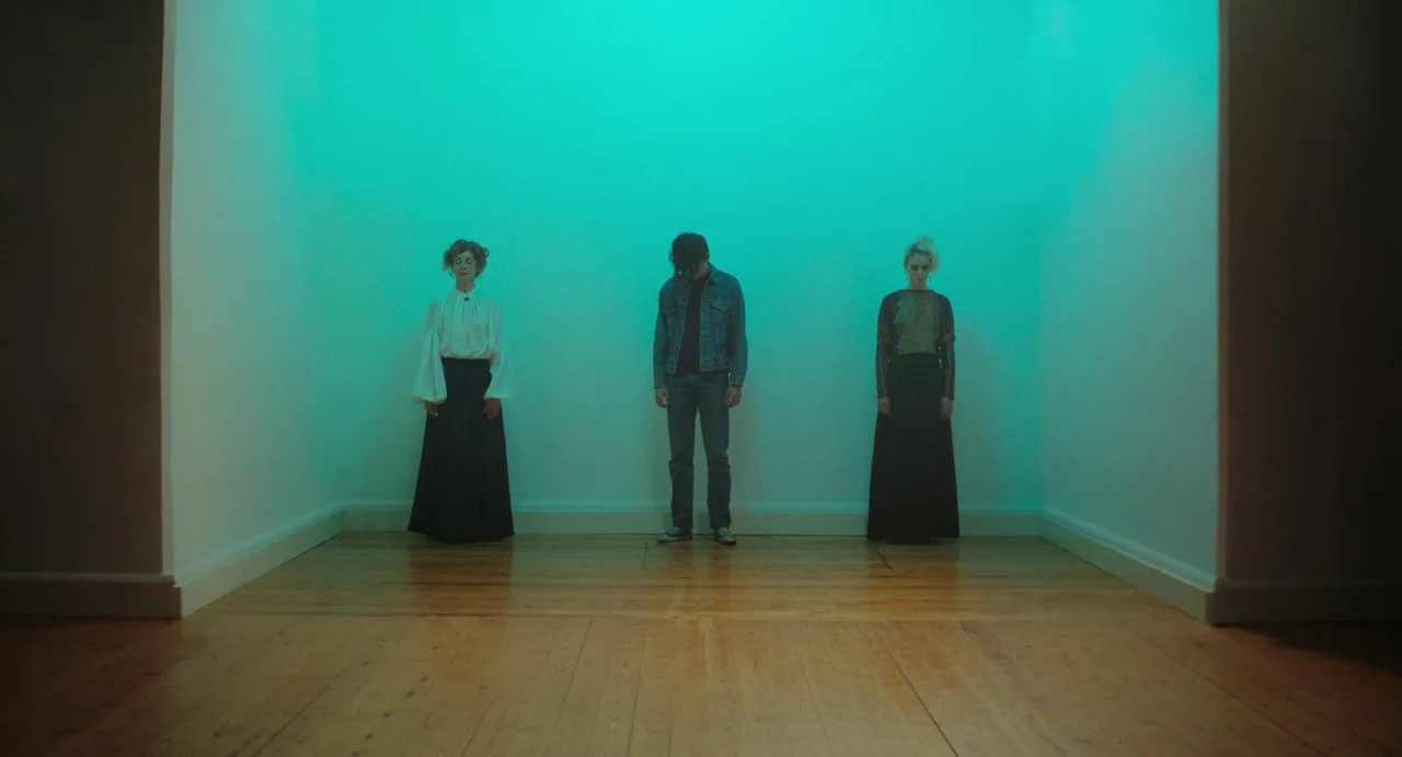 Flux Gourmet (2022) movie still: wide — Three people stand at equidistant intervals against a plain white wall in a room. The room features bare…