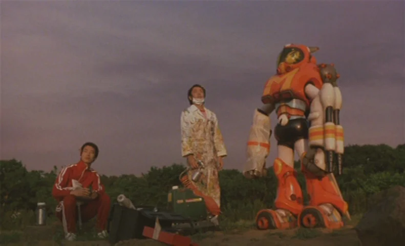 The Taste of Tea (2004) movie still: wide — An open field at dusk with trees visible in the distance under a cloudy sky. Two men stand with a…
