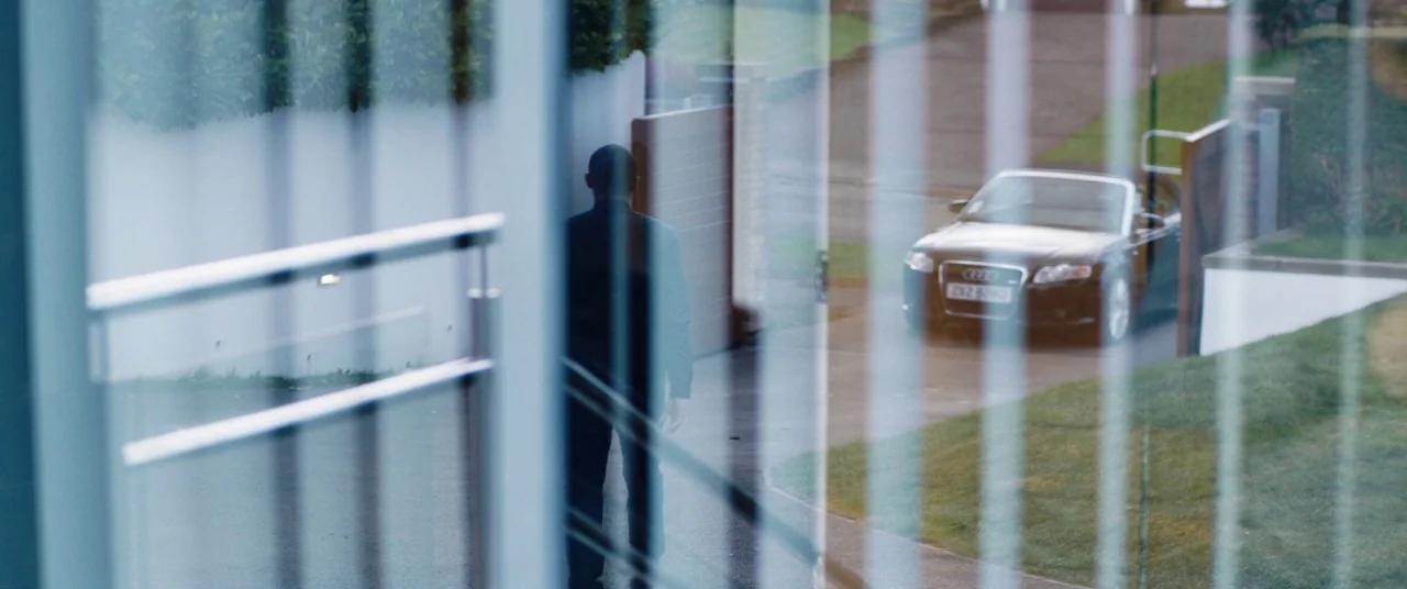 Journeyman (2017) movie still: medium — A man walks past a modern wooden gate on a paved residential driveway. A dark convertible car is parked…