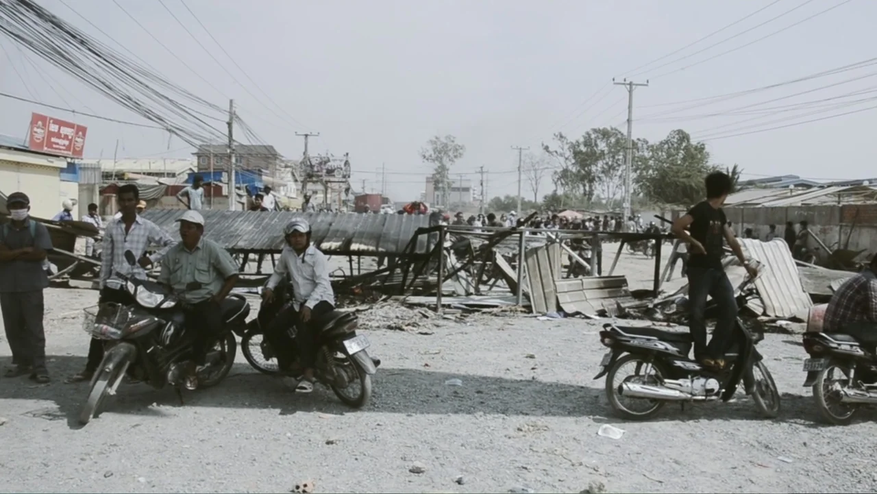 A Cambodian Spring (2018) movie still: wide — A dry, dusty lot contains overturned metal structures and several motorbikes with riders present. A…