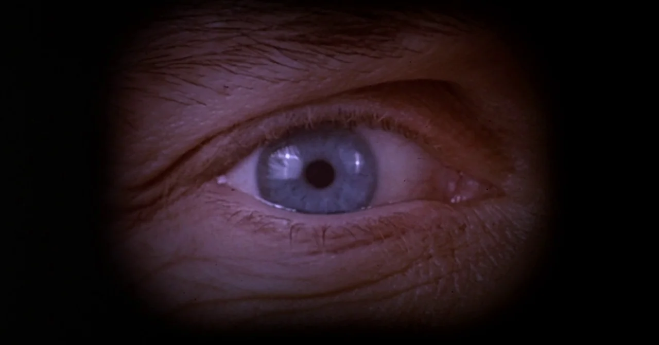 Darkman (1990) movie still: closeup — A close-up view of a blue eye looking through a circular door peephole. The surrounding eye area is visible…