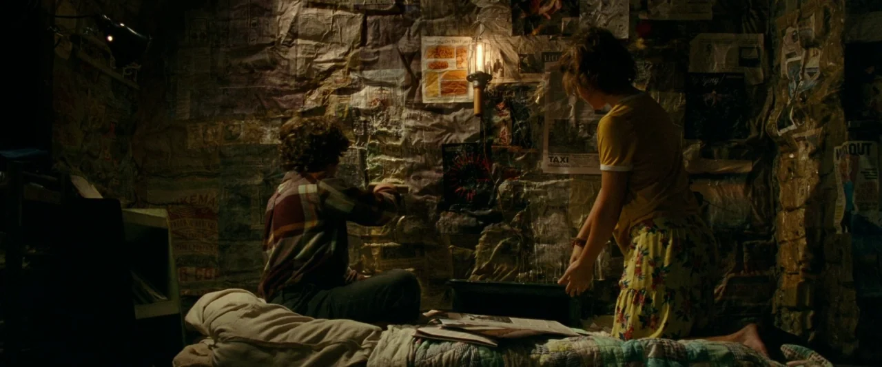 A Quiet Place (2018) movie still: medium — A bedroom wall covered in multiple layered newspaper clippings and taped documents. Two teenagers kneel…